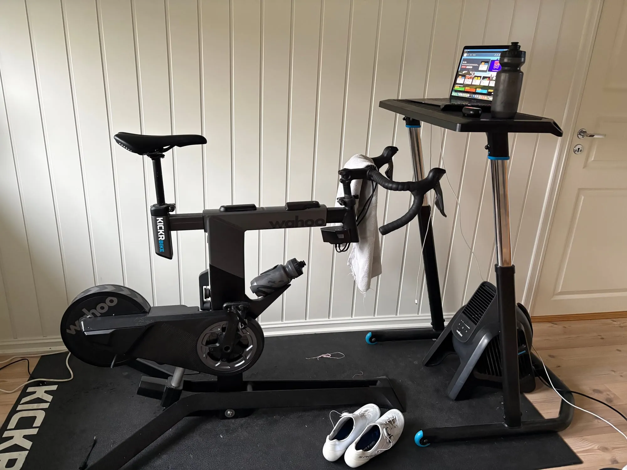 Wahoo KICKR Bike sett fra siden på en treningsmatte, med vifte foran og et høydejusterbart bord med Mac og drikkeflaske