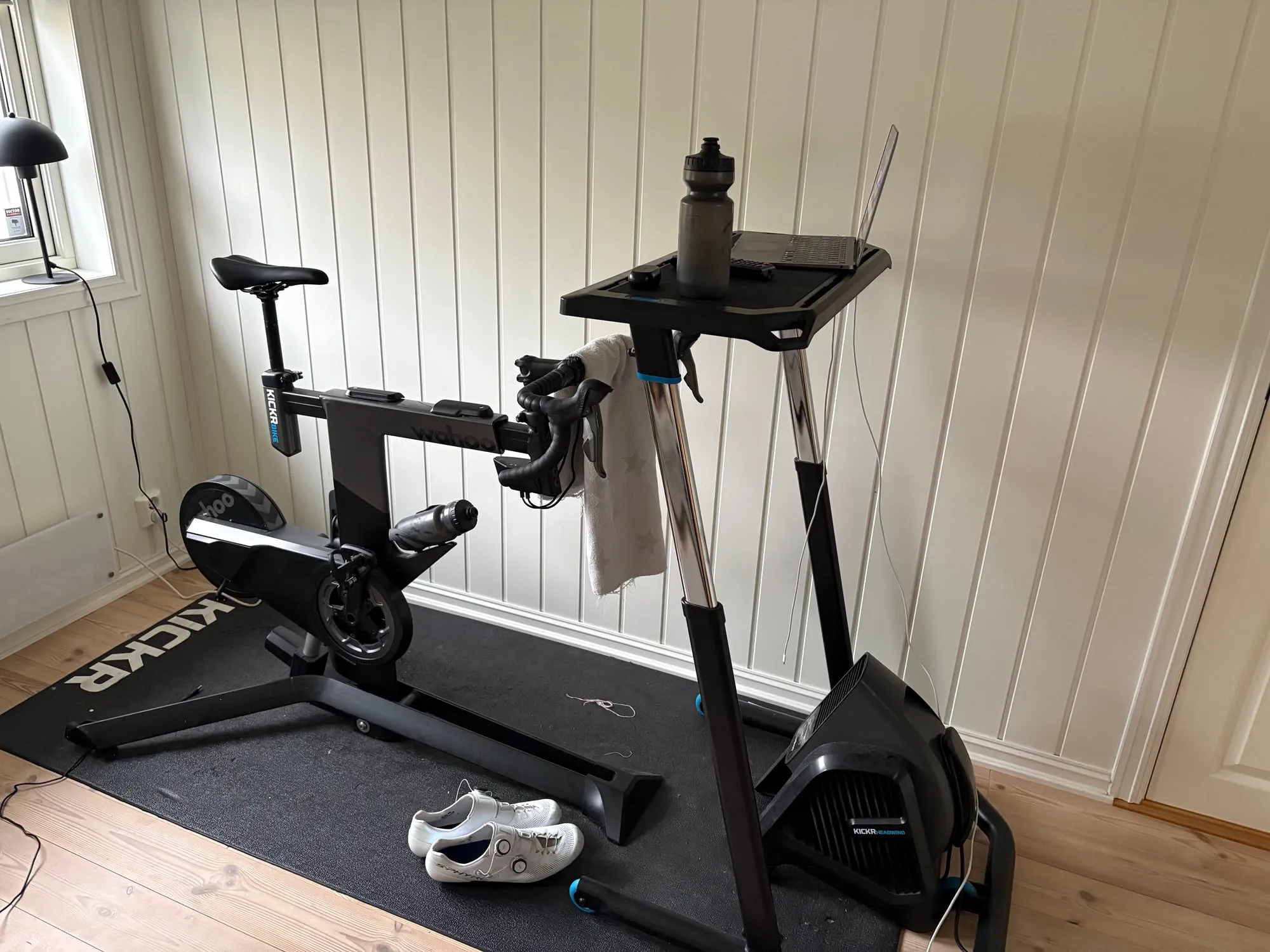 Treningshjørnet i kjelleren: Wahoo KICKR Bike på en svart matte, med vifte foran, rullebord med drikkeflaske og håndkle over styret